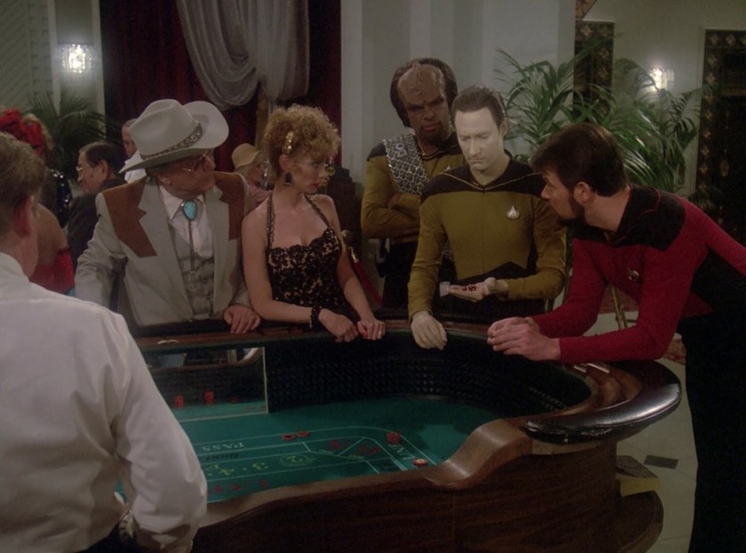 At a craps table, Commander Data looks with robo-concern at the dice in his hand. Riker, Worf, and some characters from the casino hotel watch, puzzled.