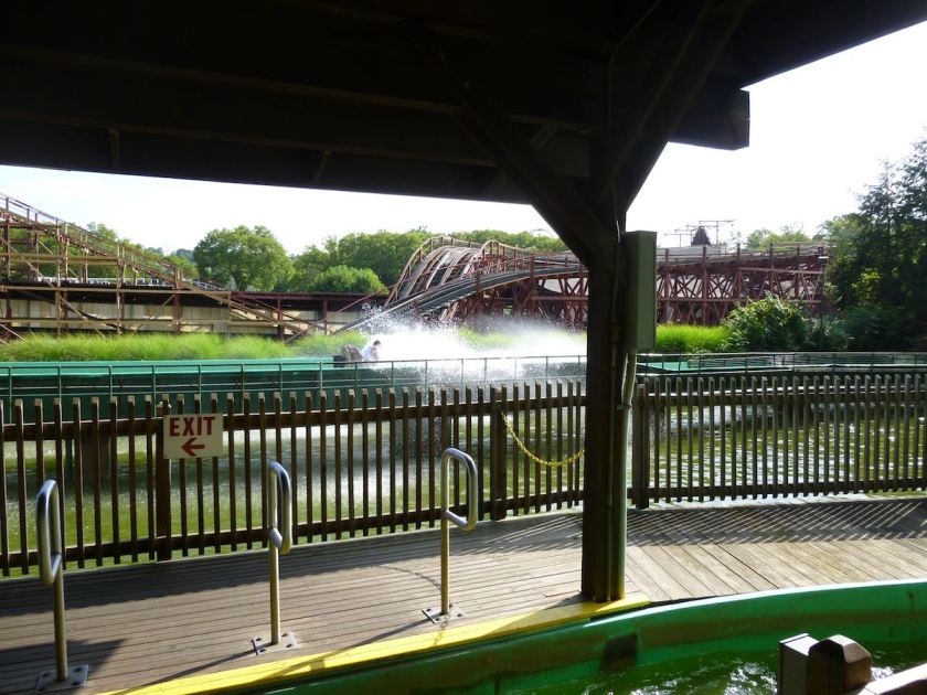 Log flume car splashing down at the bottom of its hill. In the background is the rust-red track and structure of the Racer roller coaster.