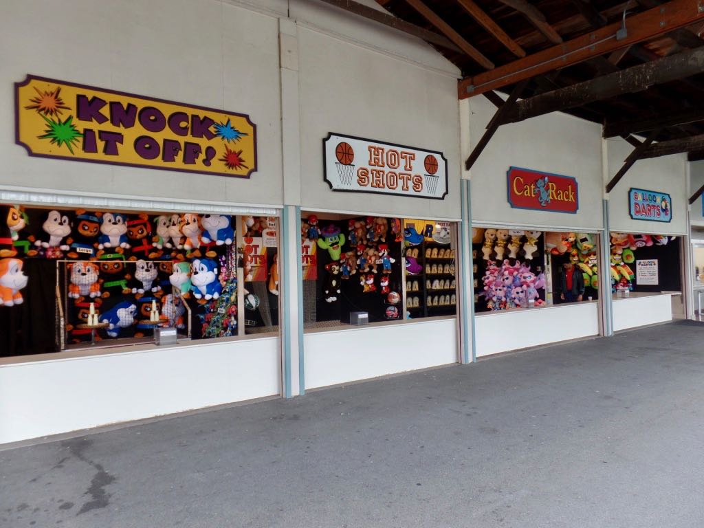 Photograph of a couple of midway games: Knock It Off (a milk-cans game), Hot Shots (basketball game), Cat Rack (knocking over cat figures), and Balloon Darts (a pop-the-balloons game). No one is playing them yet and it's not clear they're attended.